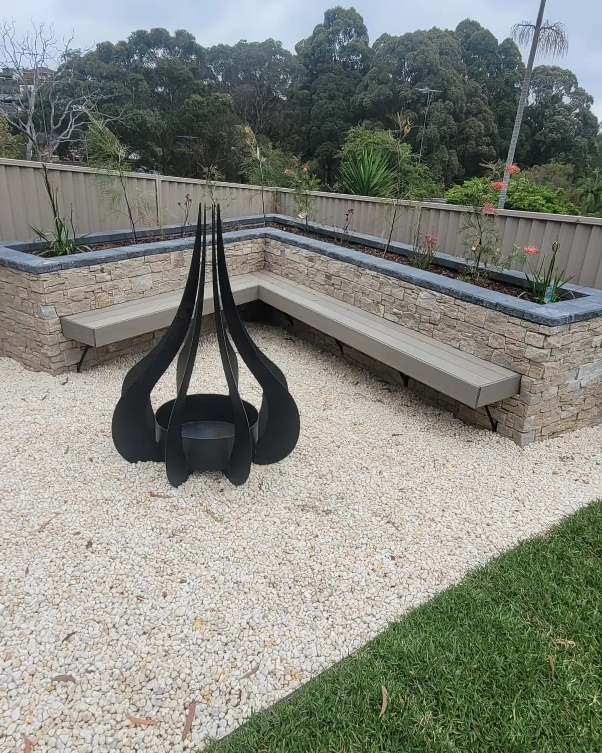 Stacked stone planter walls with a built-in bench, white gravel and a black steel fire pit